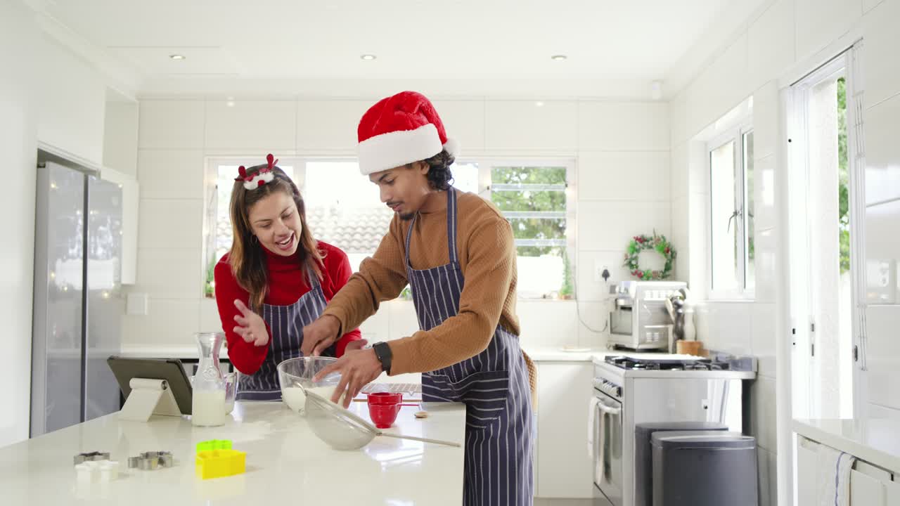 Couple cheering in aprons and antlers cracking eggs, whisking holiday batter at kitchen island
