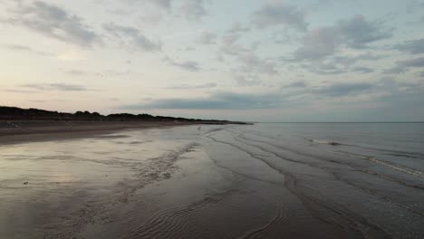 Imágenes-De-Video-Aéreas-Muestran-Una-Pintoresca-Playa-Al-Atardecer,-Donde-La-Arena-Húmeda-Y-Reflectante-Se-Encuentra-Con-El-Tranquilo-Océano-Púrpura-Y-Rosa,-Y-La-Silueta-De-La-Gente-Pasea-A-Su-Perro.