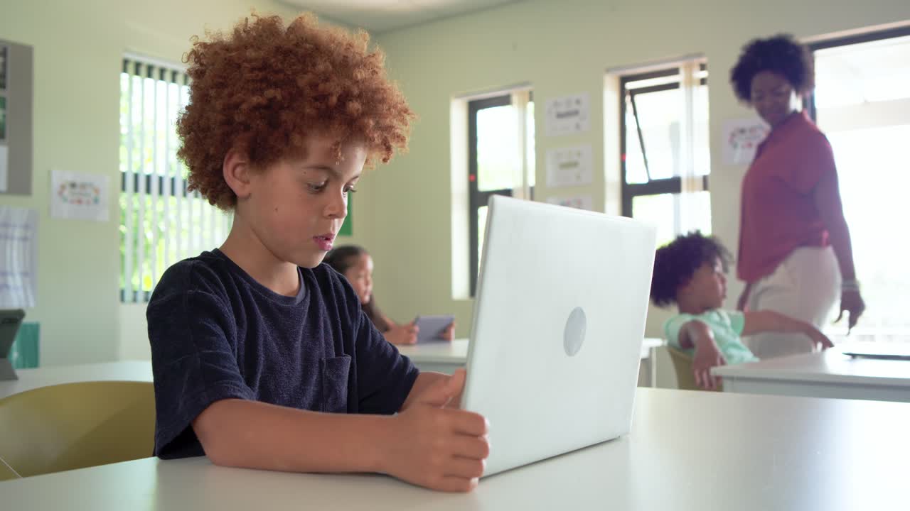 Bambino in classe, ragazzo afroamericano che posiziona un laptop bianco, svolge un compito di digitazione in classe mentre l'insegnante passa