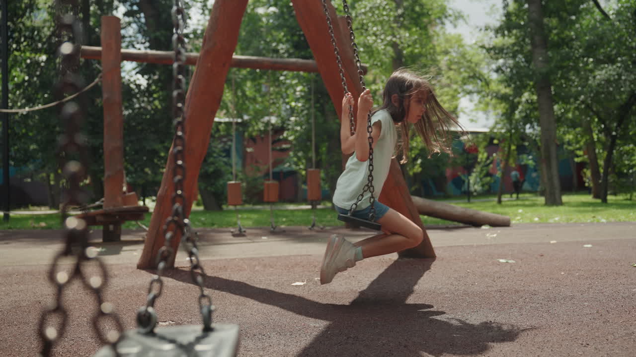 Bambina che gioca felicemente. Una bambina si gode un gioco spensierato su una superficie di sicurezza sotto il sole. Una giovane ragazza si dondola e calcia felicemente sotto la luce brillante del sole, sotto alberi frondosi, su una pavimentazione di sicurezza in gomma.