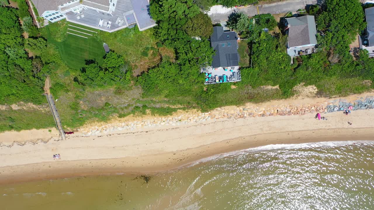Bird’s eye drone view of sandy shoreline where beach houses back onto dense greenery, with stairways leading down to the sand and turquoise waves shimmering along the coast