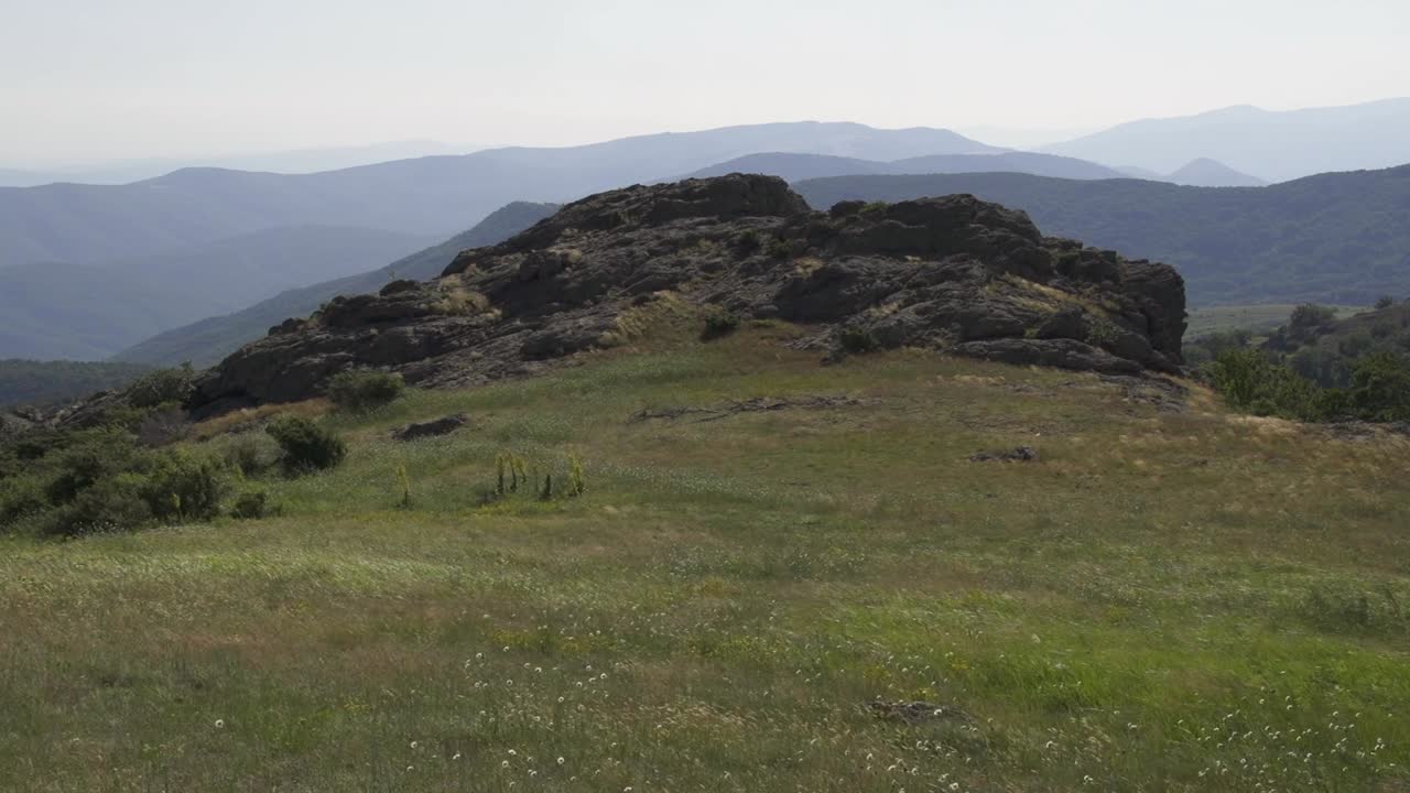 A rocky formation sits amidst a vast grassy area with distant mountains in the background. The beauty of nature on a clear day. Stara Planina next to Sliven city. Balkan Mountains