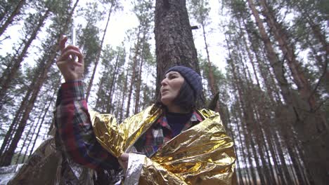 confused lost female tourist wrapped in a golden survival blanket in the forest catches a connection on her smartphone