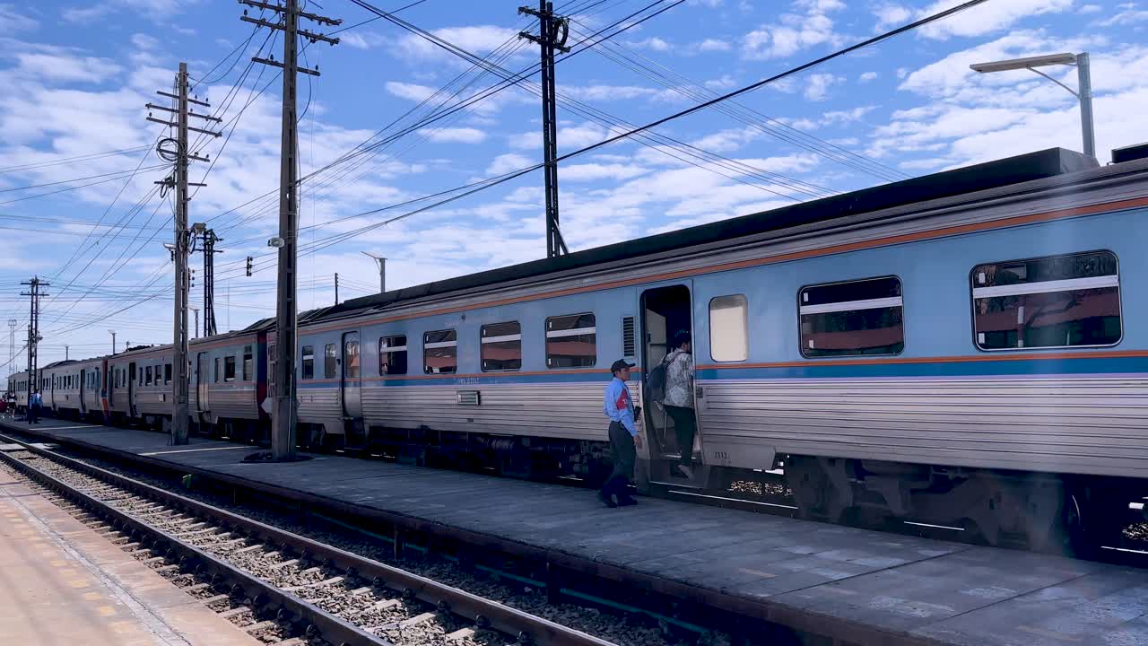 Railway guard steps onto stationary local passenger train at rural Thai station under bright daylight