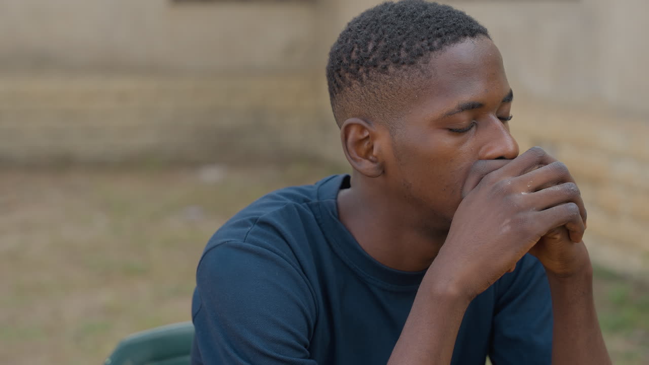 Pensive Black carpenter seated outdoors reflecting on dusty hands and fatigue, quiet yard backdrop, hands clasped near mouth, solemn expression after hard day of carpentry, authentic portrait