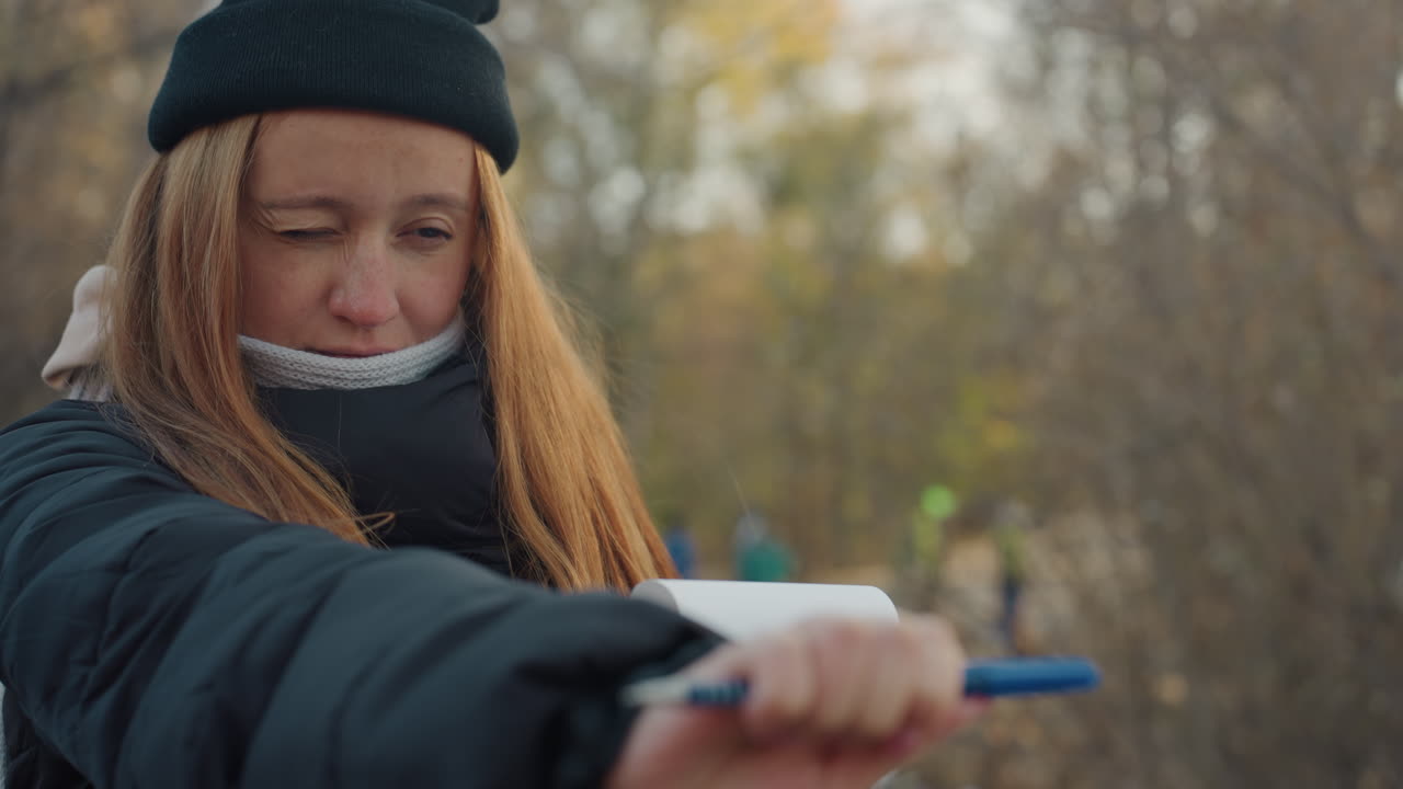 Donna che prende appunti in un parco autunnale, avvolta in cappotto e berretto, espressione concentrata mentre sfoglia pagine e annota osservazioni, sfondo di alberi spogli e foglie sparse, luce fredda, scatto spontaneo.