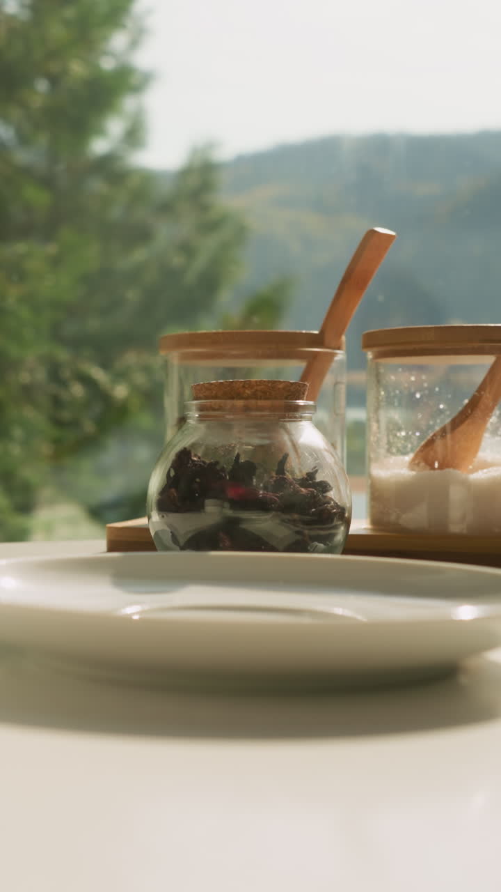 젊은 여성은 테이블에 차를 마시기 위해 접시를 놓습니다. 아내는 테이블 아침 식사를 준비합니다. 약초 차 컵이 이미 위치합니다. 양육하고 활력을 불어넣는 아침 의식.