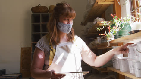 Female-caucasian-potter-wearing-face-mask-and-apron-using-digital-tablet-and-checking-pottery-stock-