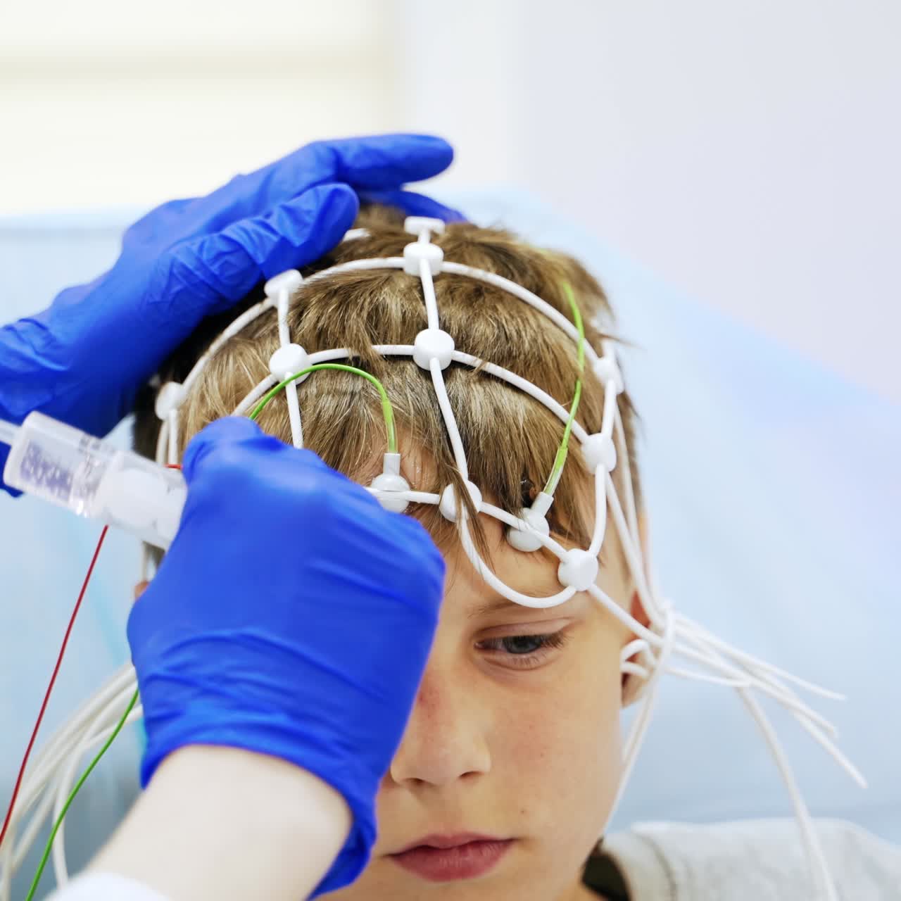 Little serious blond teenage boy with sensors for EEG examination. Close up. Gloved hands of medic attaching one more electrode to kid's head