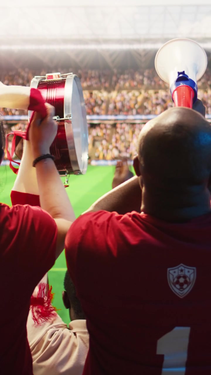 Video vertical. Aficionados al fútbol agitando bufandas durante un partido jugado en el césped de un estadio abarrotado.