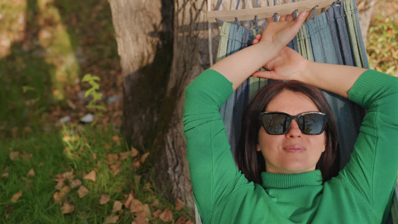 Donna in amaca in campeggio sotto un albero, occhiali da sole e maglione verde, foglie autunnali sparse; un sereno riposino di mezzogiorno e una pausa consapevole; una campeggiatrice solitaria si ricarica con un'atmosfera di benessere naturale e un accogliente ambiente esterno.