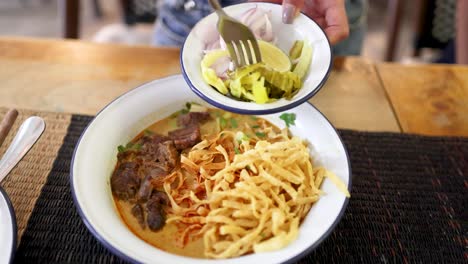 hand adds pickled vegetables, lime, and onions to a bowl of beef curry noodles in a well-lit bangkok restaurant, captured in a close-up overhead view