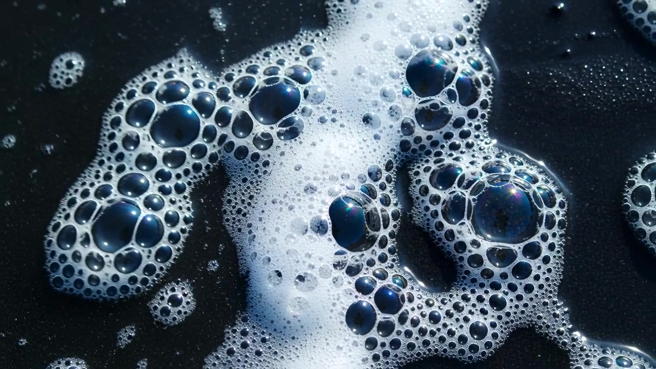 Bolle di sapone e schiuma che si spostano su un liquido scuro mentre una fascia di schiuma in arrivo ne forza la fusione.
