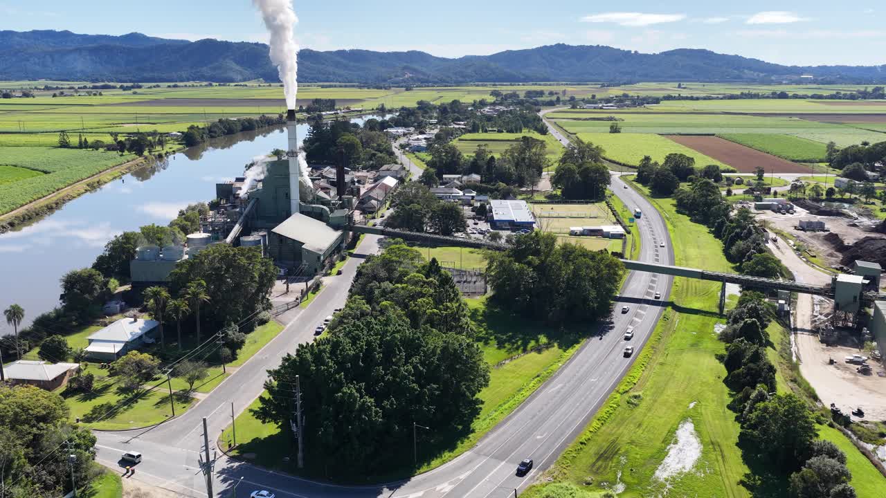 Aerial footage captures a sugar mill with smoke stacks, roads, and lush greenery in Murwillumbah, NSW, under bright daylight