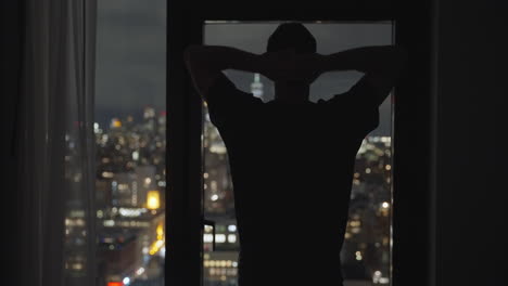 silhouette of a man leaning against a window, with his hands on his head, gazing out at the night view of manhattan