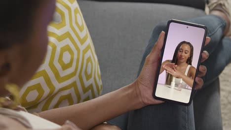 un primer plano de una mujer sentada en el sofá en casa mirando un video de demostración de un producto de belleza en un teléfono móvil
