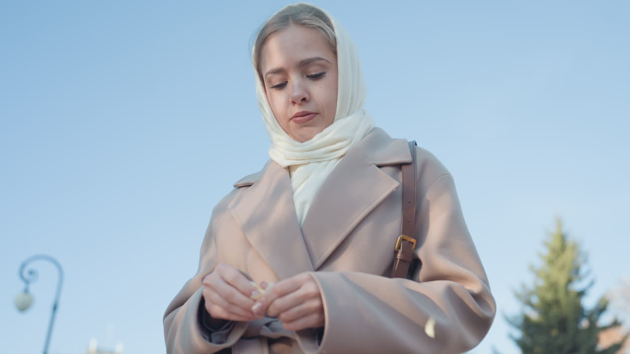 Young woman wearing headscarf outdoors beige coat adjusting ring with pensive expression, city park lampposts and evergreen trees blurred in background, cool blue sky and soft daylight, autumn