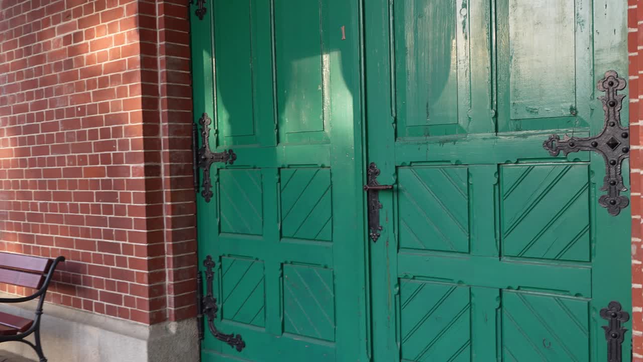 A wide view of old green wooden doors in a historic chapel in Szczecin. Benches stand in front of the red brick wall. Subtle camera movement in soft daylight.