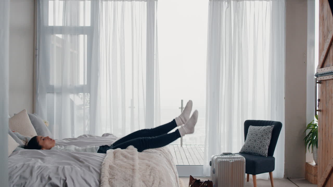 행복한 젊은 여성이 침대에 뛰어 성공적인 여행 여행 후 휴식을 취하고 호텔 방에서 독립적인 라이프 스타일 자유를 즐기고 있습니다.