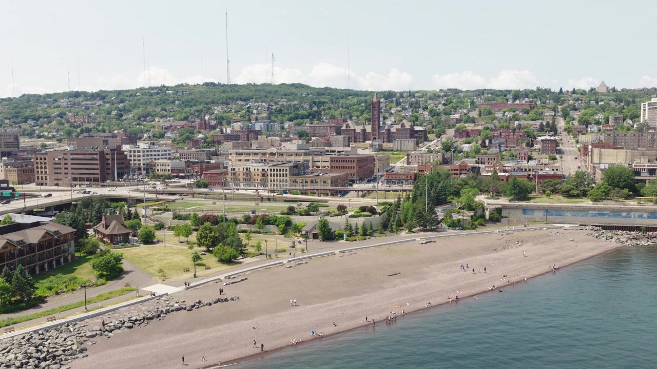Drone che si abbassa su una spiaggia sabbiosa del Lago Superiore con persone che camminano, rivelando lo skyline del centro di Duluth, Minnesota, e la collina retrostante.