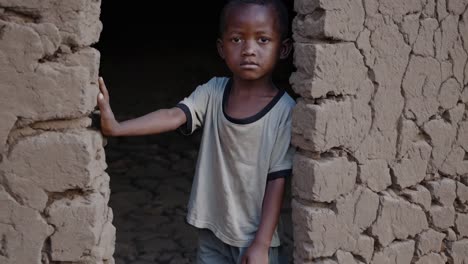 sad african child standing at house doorway, gazing directly with serious expression revealing childhood hardship and resilience in rural village environment