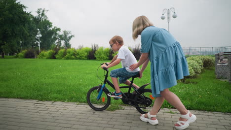 a mother gently supports her child's waist for balance while he learns to ride a bicycle close to a vibrant green garden