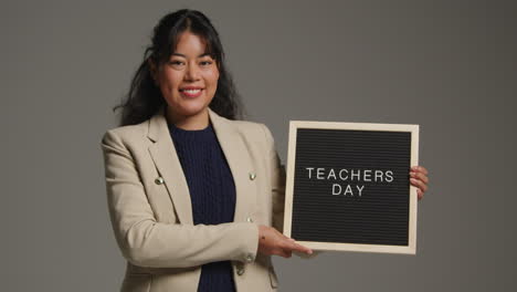 studio portrait of female teacher standing against grey background holding up notice board reading teachers day