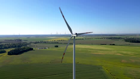 Nahaufnahme-Einer-Windkraftanlage-Mit-Anderen-Im-Hintergrund,-Rapsfeld-Und-Blauem-Himmel,-Erneuerbare-Energiequellen