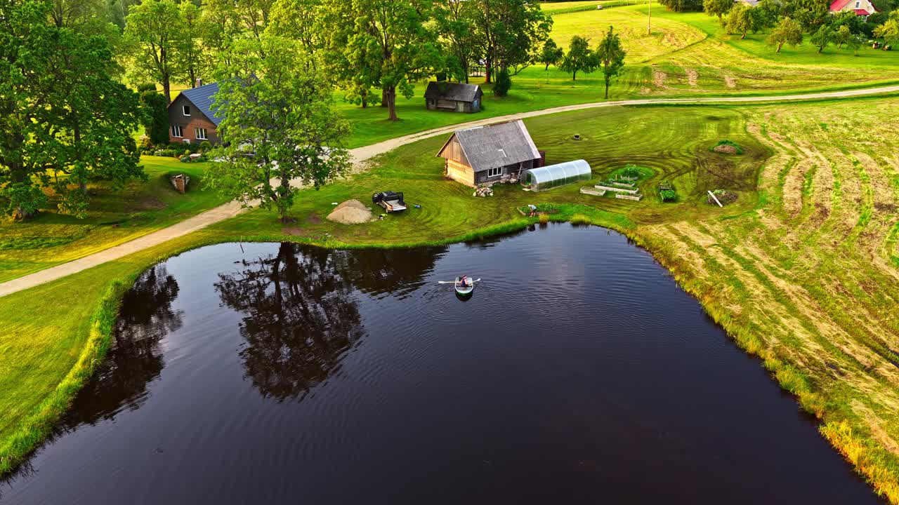 A person in a small boat floats on a calm lake beside a house, seen from above
