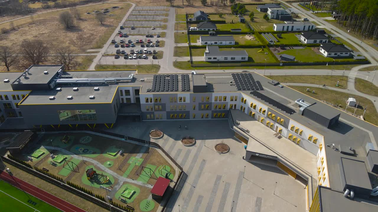 Aerial drone footage of Laagri Möldre maja highschool during a sunny autumn or spring day. The school is large and modern and has a football or a soccer stadium in front of it. Cars parked in the back