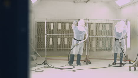 sliding shot of 2 workers wearing respirators and coating manufactured parts inside a paint spray booth
