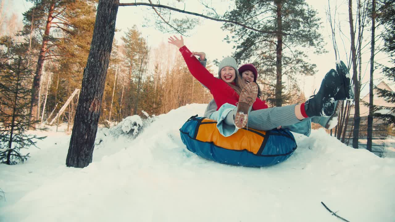 dos multiétnicas emocionadas felices y hermosas amigas mujeres sonriendo en trineo en la pendiente de nieve a la cámara, diversión de invierno en cámara lenta.