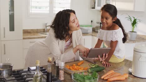 hispanic mother teaching daughter sitting on countertop cooking, looking at tablet