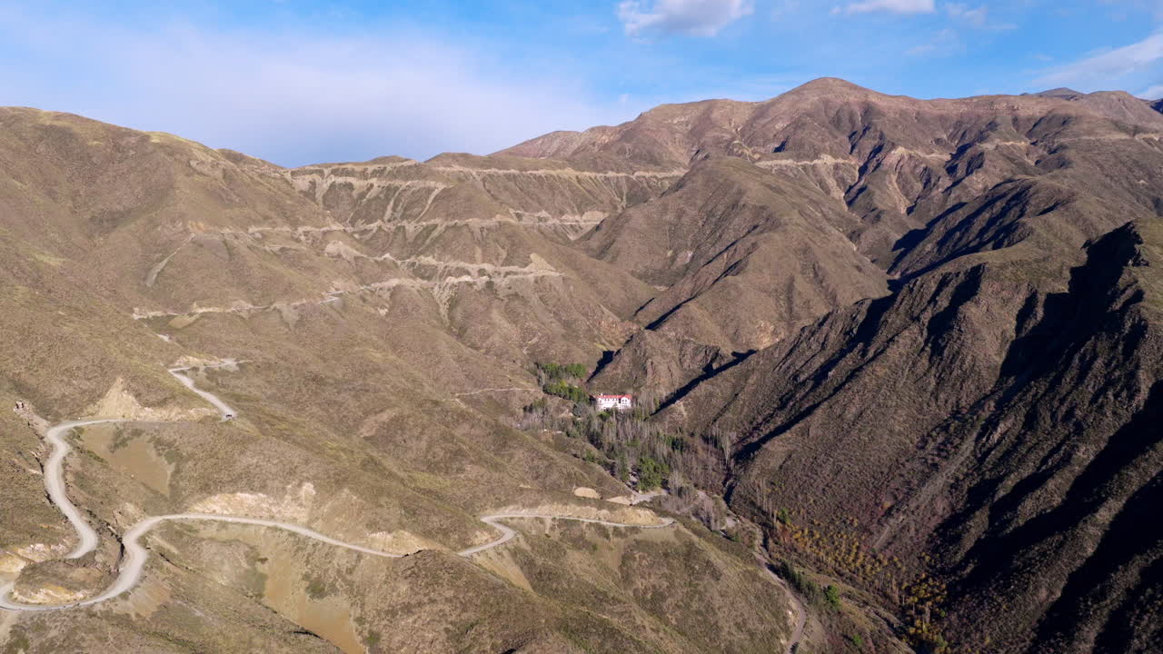 Slow panoramic drone movement reveals winding Caracoles de Villavicencio road tracing arid Andean slopes, gradually exposing Gran Hotel Villavicencio surrounded by sparse forest, Mendoza, Argentina