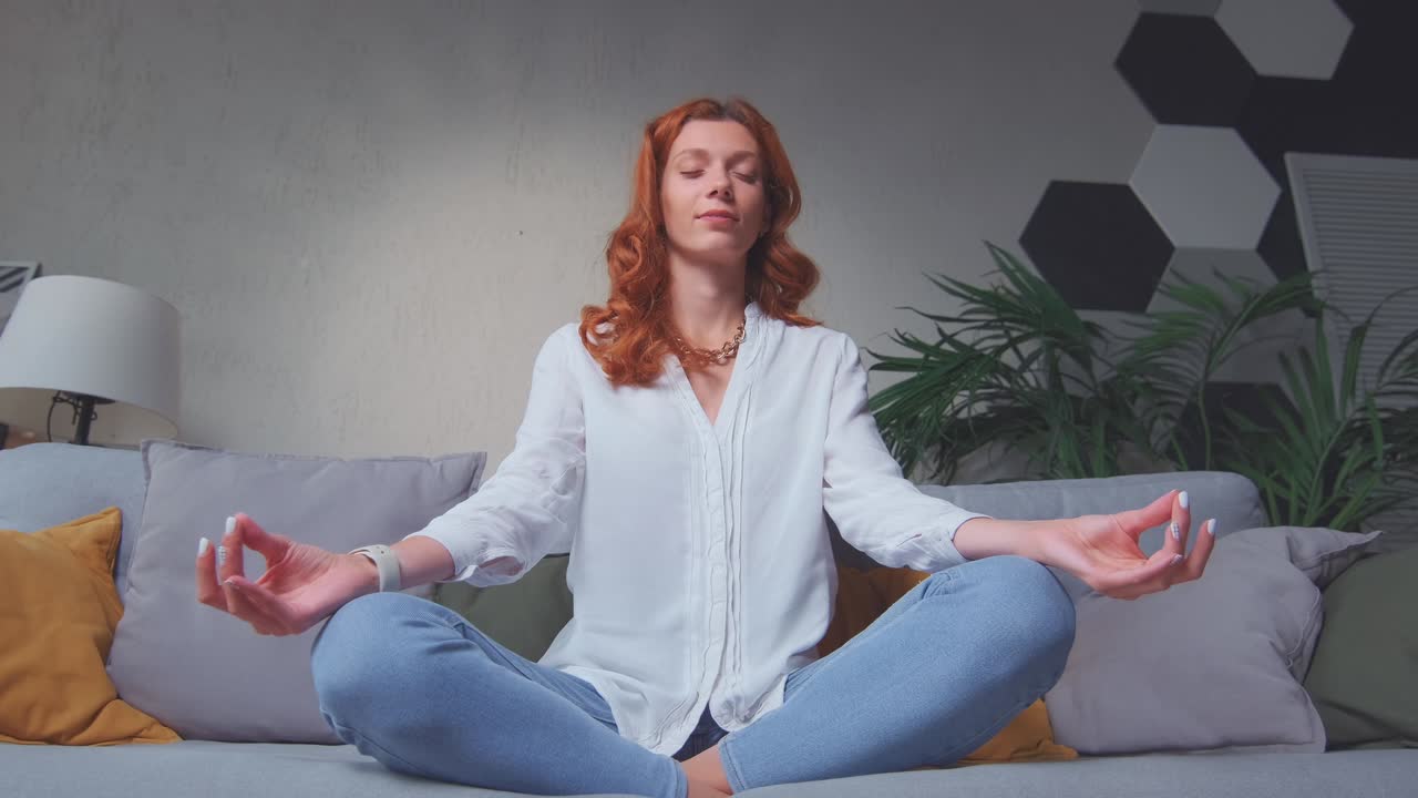 Young caucasian woman doing yoga sits on sofa in lotus position meditation