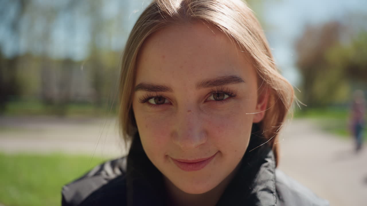 햇살이 비치는 공원에서 카메라를 향해 미소 짓는 백인 인플루언서의 클로즈업 사진이에요. 은은한 보케, 캐주얼한 재킷, 부드러운 바람, 따뜻한 색조가 진솔한 라이프스타일 콘텐츠와 친근한 개성을 전달해요.