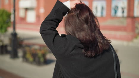 close-up back view of a young lady with brown hair in a black coat adjusting her head, showing off three rings on her left hand, on a sunny urban street