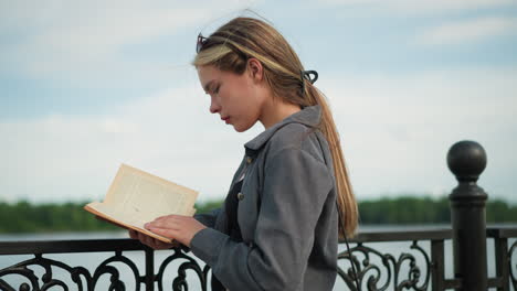 lady in grey clothing rests her hand on an iron fence while placing her left hand on the pages of her book to keep them from blowing in the wind, the wind softly blows her hair