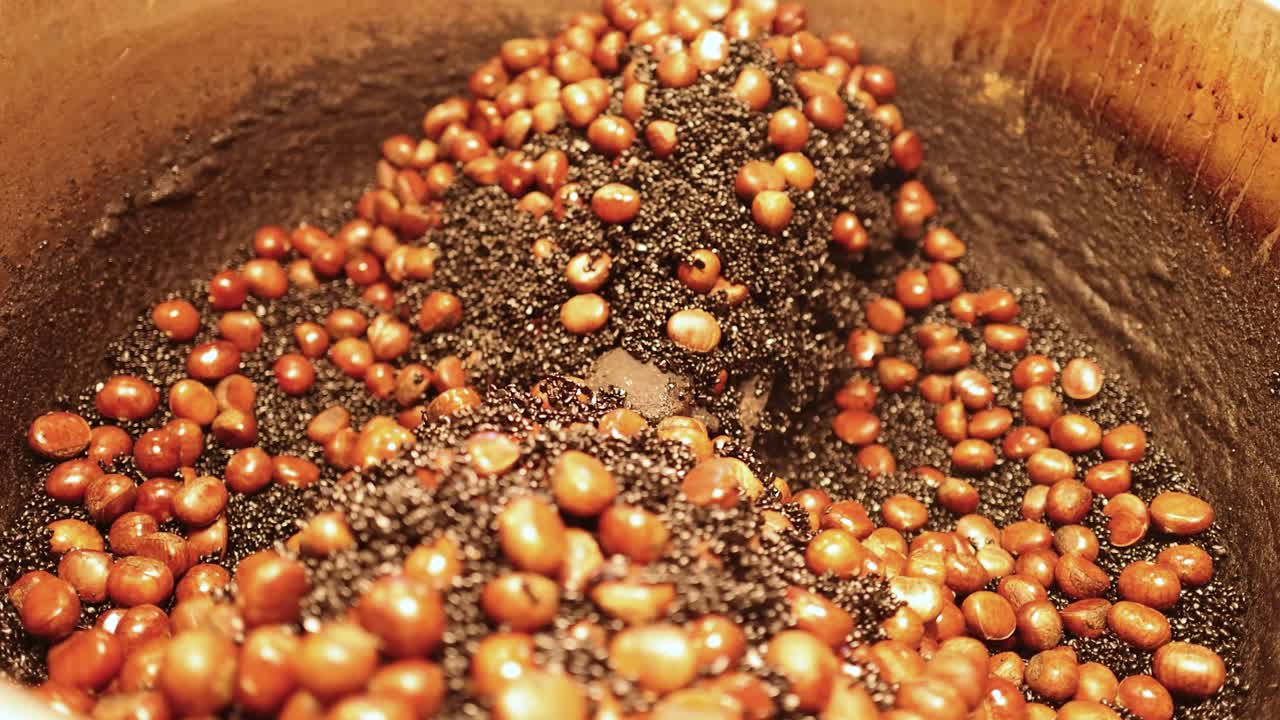 Chestnuts tumble in hot black sand inside a heated drum, glowing under warm street lights