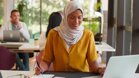 mature businesswoman wearing headscarf working at desk in office writing in notebook