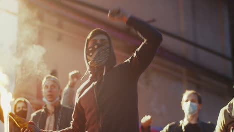 homem caucasiano com lenço no rosto gritando com os braços para cima e segurando coquetel molotov em um protesto com grupo multiétnico de pessoas na rua