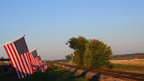 a view of a line of gently waving american flag on a fence by farmlands by a single railroad track in late afternoon