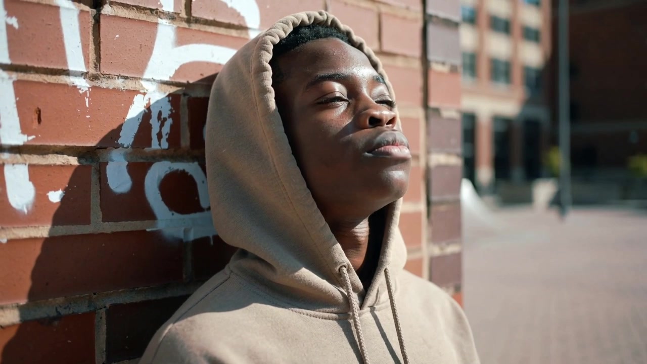 Adolescente in felpa con cappuccio beige che si appoggia, si inclina verso la luce del sole e chiude gli occhi in una piazza urbana, con un muro graffitato.