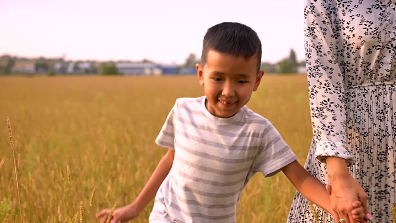 Anak laki-laki Asia yang manis berjalan selangkah demi selangkah mengikuti ibunya yang penyayang sambil memegang tangannya di ladang gandum