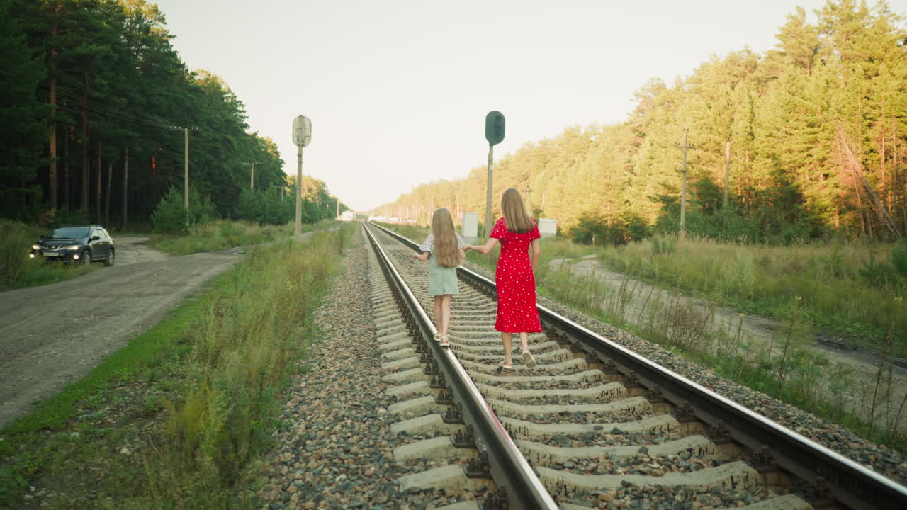 back view of woman holding child hand walking on railroad track, background features parked car on dirt road, signal posts and rural landscape with forest trees under warm natural sunlight