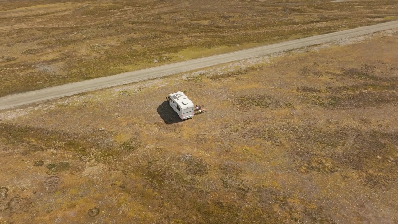 Ripresa cinematografica con drone in orbita discendente che si avvicina a un camper parcheggiato accanto a un fiume glaciale in una suggestiva valle di montagna, rivelando una remota destinazione di viaggio all'aperto.