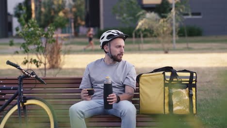 Tired-Food-Delivery-Man-Seated-On-A-Bench-Next-To-His-Thermal-Backpack-During-His-Break-Drinking-A-Cup-Of-Tea-And-Thinking-About-His-Stuff