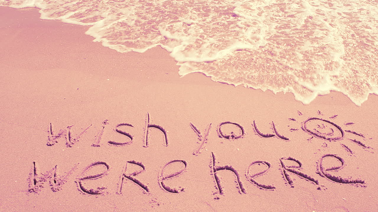 Testo concettuale 'wish you were here' scritto a mano sulla sabbia di una spiaggia.