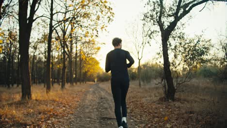 Rear-view-of-a-sporty-guy-in-a-black-sports-uniform-runs-along-an-earthen-path-in-the-autumn-forest-at-sunrise-in-the-morning