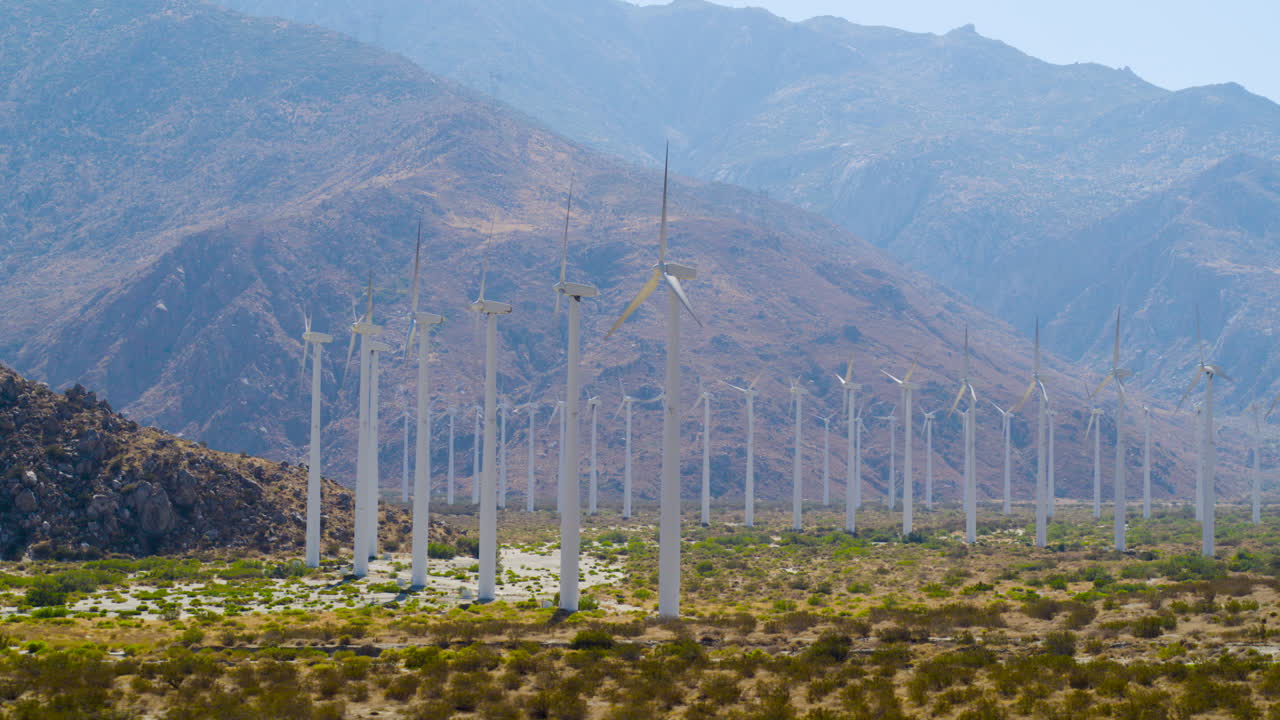 Palm Springs windmills in rows spinning with the rugged San Jacinto Mountains rising behind them in California under a clear blue sky on a bright, sunny day. Cinematic aerial shot of valley